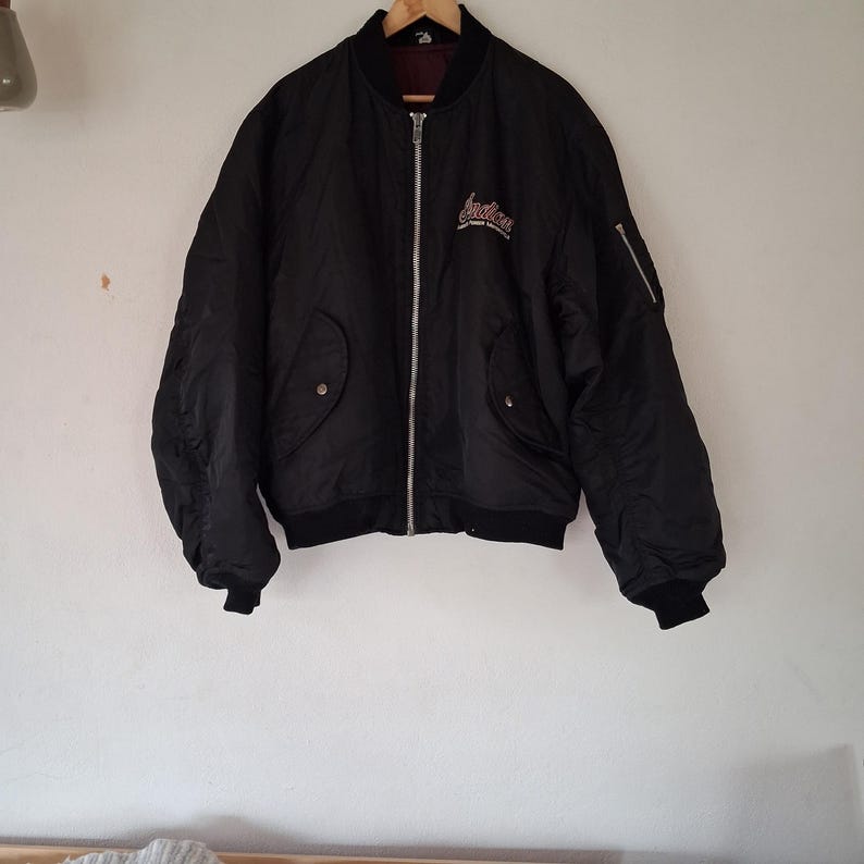 May include: A black bomber jacket featuring a silver front zipper, ribbed cuffs and hem, and two front pockets with snap closures. The chest displays white and red embroidered text that reads Indian, with a utility pocket on the left sleeve.