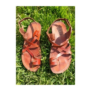 May include: A pair of brown leather sandals with a toe loop and crisscross straps. The sandals have adjustable ankle straps and are set against a backdrop of green grass.