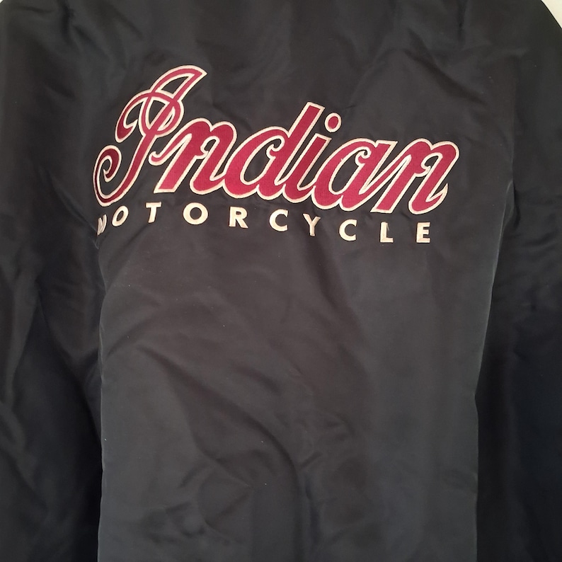 May include: A black jacket featuring the text Indian MOTORCYCLE printed in a large, stylized red font with a white outline across the upper back. The garment is made of a smooth, dark fabric with a subtle sheen and a visible texture.