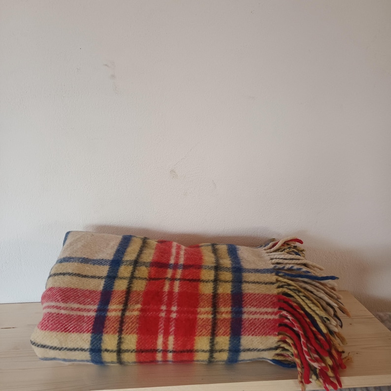May include: A folded blanket with a plaid pattern in red, blue, yellow, and beige. The blanket has a fringe on one end. The blanket is resting on a wooden surface.