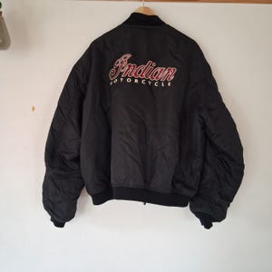 May include: A black bomber jacket featuring the text Indian Motorcycle embroidered in red and white on the back. The jacket has a ribbed black collar, cuffs, and hem, with long sleeves and a smooth, solid black nylon-style outer shell fabric.