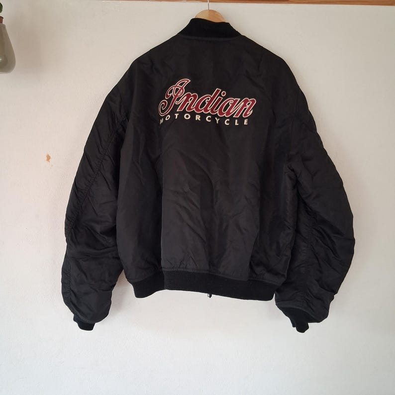 May include: A black bomber jacket featuring the text Indian Motorcycle embroidered in red and white on the back. The jacket has a ribbed black collar, cuffs, and hem, with long sleeves and a smooth, solid black nylon-style outer shell fabric.