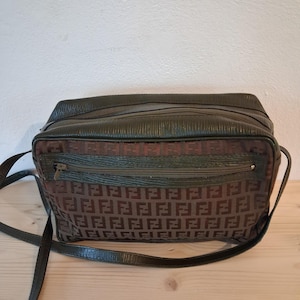 Puede incluir: Bolso de hombro rectangular en marrón y verde oscuro. El bolso presenta un patrón marrón repetido en la parte delantera y una parte superior y ribete de cuero verde oscuro. Tiene una correa larga y ajustable y un bolsillo frontal con cremallera.