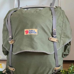 Puede incluir: Una mochila Fjällräven de color verde oliva con un marco externo de metal. La bolsa cuenta con una solapa superior grande, correas grises ajustables y detalles de cuero color canela claro. El parche rectangular del logo Fjällräven está centrado.