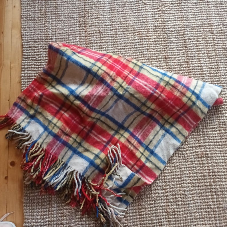May include: A folded plaid blanket with red, white, blue, and yellow stripes. The blanket has a fringed edge with red, yellow, and blue tassels. The blanket is made of a soft, woven material.