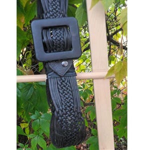 Puede incluir: Cintur&oacute;n de cuero negro con una hebilla cuadrada grande y detalles trenzados. El cintur&oacute;n cuelga sobre un fondo de hojas verdes y una estructura de madera.