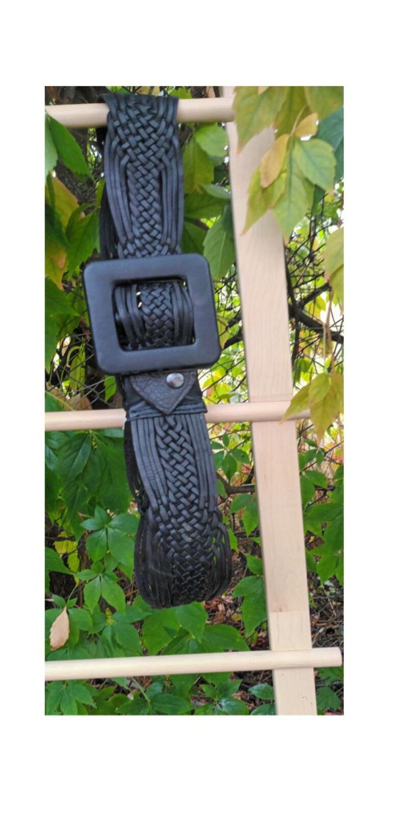 Puede incluir: Cintur&oacute;n de cuero negro con una hebilla cuadrada grande y detalles trenzados. El cintur&oacute;n cuelga sobre un fondo de hojas verdes y una estructura de madera.