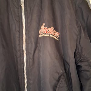 May include: A black zip-up jacket featuring a silver-toned metal zipper closure. The chest area displays the embroidered text Indian America's Pioneer Motorcycle in red and white lettering, positioned on the left side of the garment.