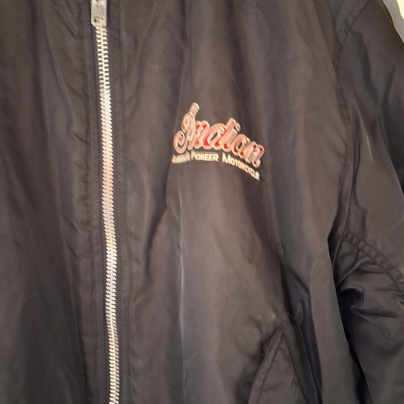 May include: A black zip-up jacket featuring a silver-toned metal zipper closure. The chest area displays the embroidered text Indian America's Pioneer Motorcycle in red and white lettering, positioned on the left side of the garment.