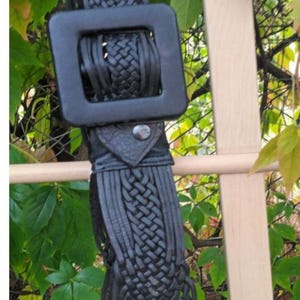 Puede incluir: Cintur&oacute;n de cuero negro con una hebilla cuadrada grande. El cintur&oacute;n presenta un dise&ntilde;o tejido y flecos. El cintur&oacute;n cuelga de una estructura de madera con follaje verde en el fondo.