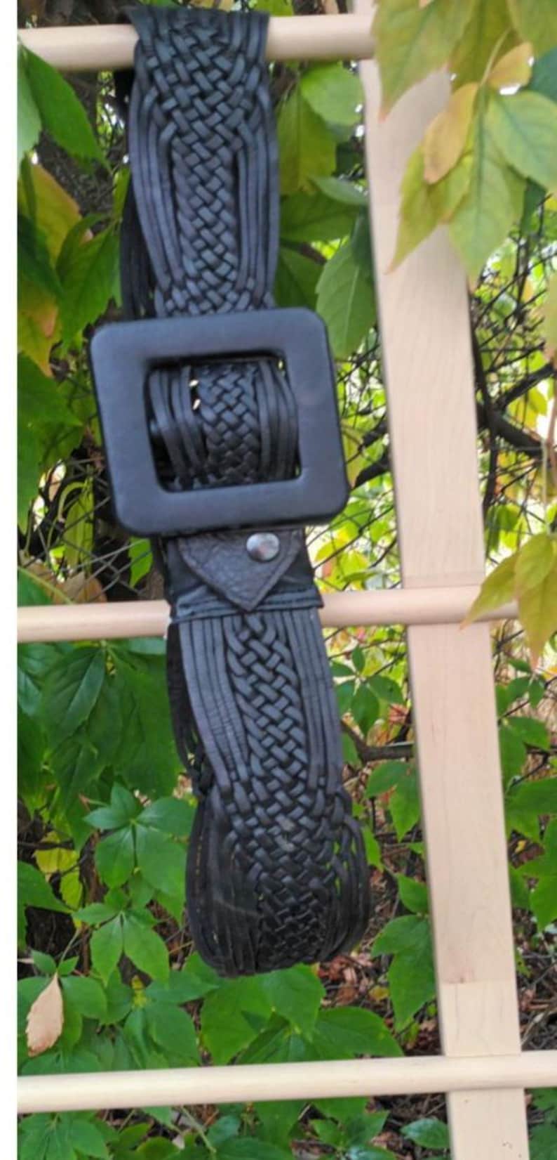 Puede incluir: Cintur&oacute;n de cuero negro con una hebilla cuadrada grande. El cintur&oacute;n presenta un dise&ntilde;o tejido y flecos. El cintur&oacute;n cuelga de una estructura de madera con follaje verde en el fondo.