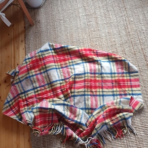 May include: A red, blue, and yellow plaid blanket with a cream background. The blanket has a fringed edge and is folded on a woven rug. The blanket is made of a soft material.
