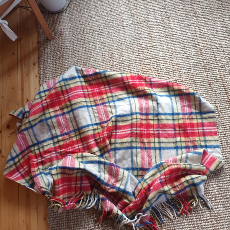 May include: A red, blue, and yellow plaid blanket with a cream background. The blanket has a fringed edge and is folded on a woven rug. The blanket is made of a soft material.