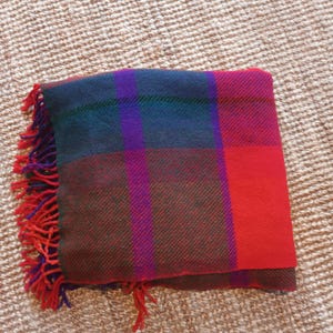 May include: A folded plaid blanket with a red, purple, green, and dark red pattern. The blanket has a fringe of red and purple threads along one edge. The blanket is made of a woven material.