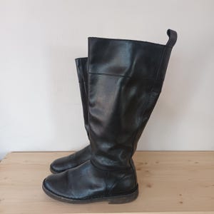 May include: A pair of black knee-high boots. The boots are made of a smooth, leather-like material. They have a rounded toe and a low, flat sole. The boots are standing on a wooden surface.