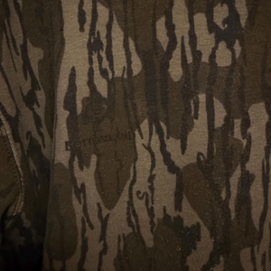 May include: Close-up of a camouflage pattern in shades of brown, olive green, and black. The fabric has a textured appearance with the word "BOTTOMLAND" faintly visible.