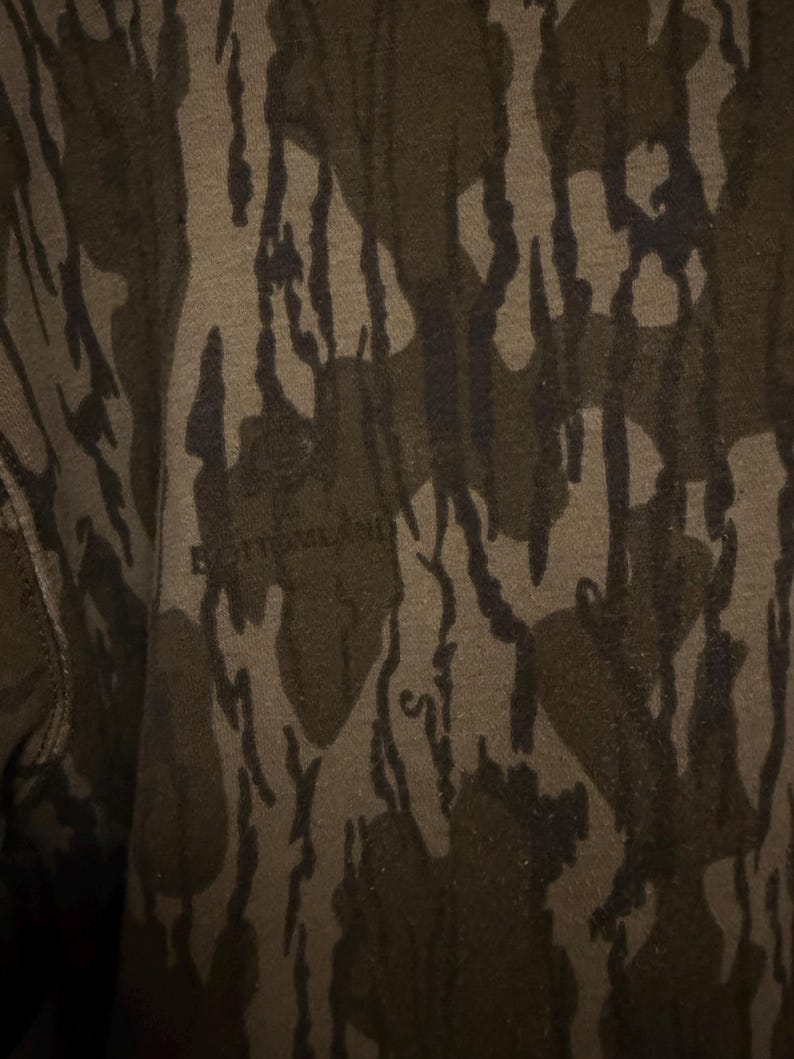 May include: Close-up of a camouflage pattern in shades of brown, olive green, and black. The fabric has a textured appearance with the word "BOTTOMLAND" faintly visible.