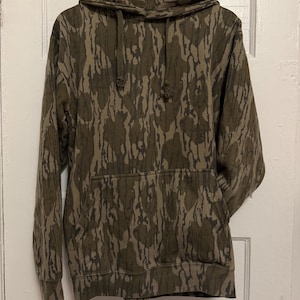 May include: A camouflage-patterned hoodie in shades of brown and olive green hangs on a wooden hanger. The sweatshirt features a front pocket and drawstrings on the hood. The fabric has a textured appearance.