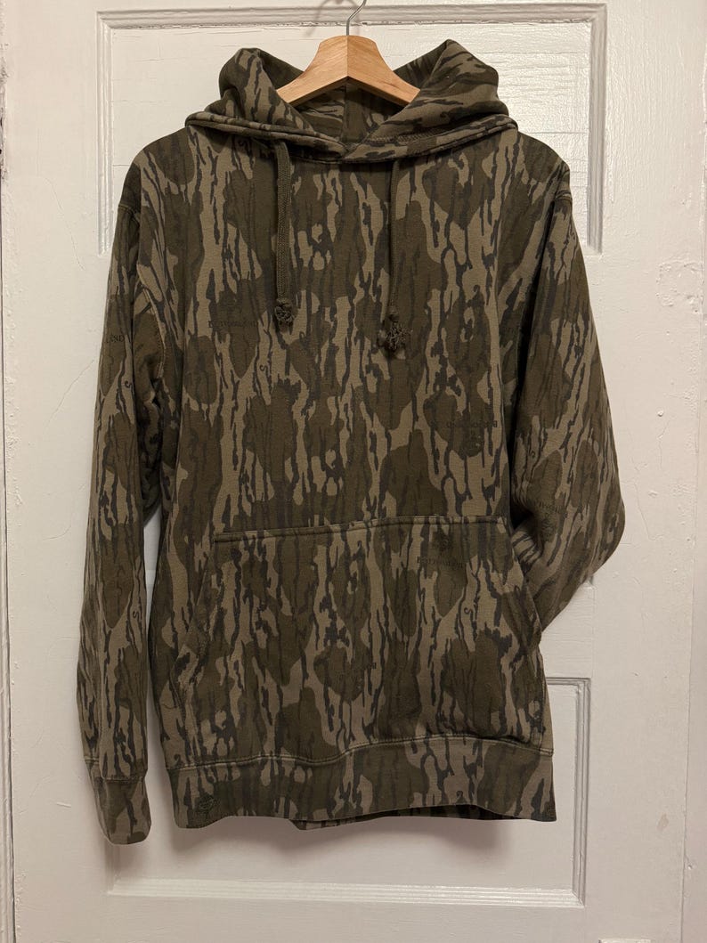 May include: A camouflage-patterned hoodie in shades of brown and olive green hangs on a wooden hanger. The sweatshirt features a front pocket and drawstrings on the hood. The fabric has a textured appearance.