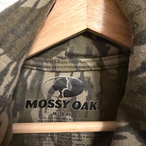 May include: A close-up of a camouflage-patterned hoodie with the "MOSSY OAK" logo. The tag indicates the size is M (38-40) and the fabric composition is 55% cotton and 45% polyester. The hoodie is hanging on a wooden hanger.