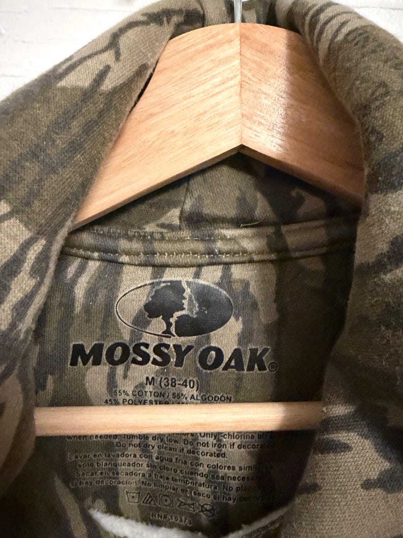 May include: A close-up of a camouflage-patterned hoodie with the "MOSSY OAK" logo. The tag indicates the size is M (38-40) and the fabric composition is 55% cotton and 45% polyester. The hoodie is hanging on a wooden hanger.