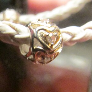 May include: A gold-colored charm with heart-shaped cutouts and a small gemstone in the center. The charm is on a braided, off-white cord. The charm is likely part of a bracelet or necklace.