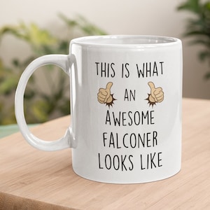 May include: White ceramic coffee mug with black text that reads "This is what an awesome falconer looks like" with two thumbs up emojis on either side of the text.