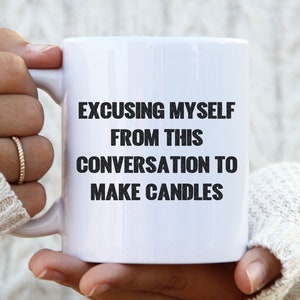 Könnte beinhalten: Weiße Keramik-Tasse mit dem Text "EXCUSING MYSELF FROM THIS CONVERSATION TO MAKE CANDLES" in Schwarz. Die Tasse wird von einer Person gehalten, die einen weißen Zopfstrickpullover trägt. Die Tasse hat eine Standardgröße.