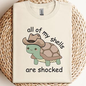 May include: Cream-colored t-shirt featuring a cartoon turtle wearing a cowboy hat. The turtle has a green body and a brown shell. The text "all of my shells are shocked" is printed in black.