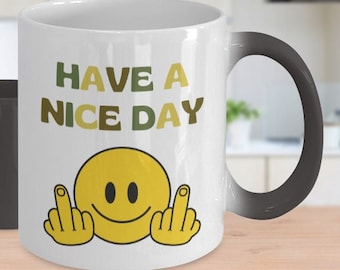 May include: A white ceramic mug with a black handle and a colour-changing design. The mug displays the text "HAVE A NICE DAY" with a yellow smiley face giving the middle finger. The mug turns black when cold.