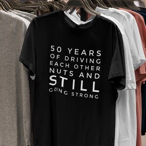 Może przedstawiać: Czarny t-shirt z białym tekstem, kt&oacute;ry brzmi "50 lat jazdy na siebie nawzajem orzech&oacute;w i nadal silny". Koszulka wisi na wieszaku z innymi koszulkami w sklepie odzieżowym.