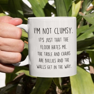 Peut inclure: Une tasse en céramique blanche avec un motif de texte noir qui dit "Je ne suis pas maladroit. C'est juste que le sol me déteste. La table et les chaises sont des brutes et les murs se mettent en travers du chemin."