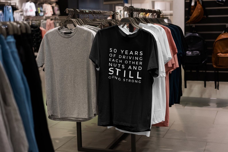 Może przedstawiać: Czarny t-shirt z białym tekstem, kt&oacute;ry brzmi "50 lat jazdy na siebie nawzajem orzech&oacute;w i nadal silny". Koszulka wisi na wieszaku z innymi koszulkami w sklepie odzieżowym.