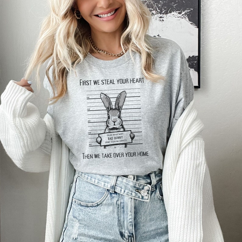 K&ouml;nnte beinhalten: Graues T-Shirt mit einem Grafikdruck eines Kaninchens in einer Polizeiaufstellung. Der Text auf dem Shirt lautet "First we steal your heart, then we take over your home".