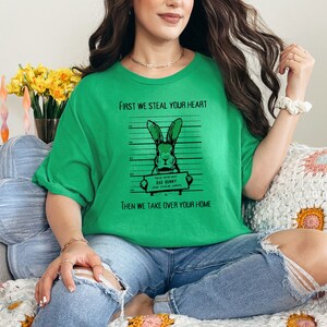 K&ouml;nnte beinhalten: Gr&uuml;nes T-Shirt mit einem Grafikdruck eines Kaninchens in einer Polizeiaufstellung. Der Text auf dem Shirt lautet "First we steal your heart, then we take over your home".