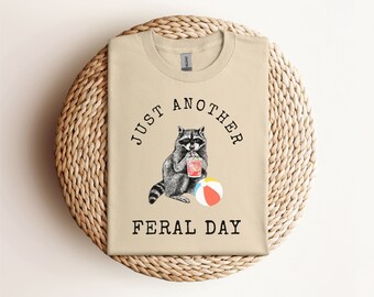 Puede incluir: Una camiseta de color beige con el texto "Just Another Feral Day" impreso en ella. Un mapache de dibujos animados sostiene un cóctel y una pelota de playa.