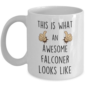 May include: White ceramic coffee mug with a black graphic that says "This is what an awesome falconer looks like" with two thumbs up emojis on either side of the text.