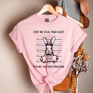 K&ouml;nnte beinhalten: Rosafarbenes T-Shirt mit einer schwarz-wei&szlig;en Illustration eines Kaninchens, das ein Schild mit der Aufschrift "Police Department Bad Bunny Crime Stealing Carrots" h&auml;lt. Der Text auf dem T-Shirt lautet "First we steal your heart then we take over your home".