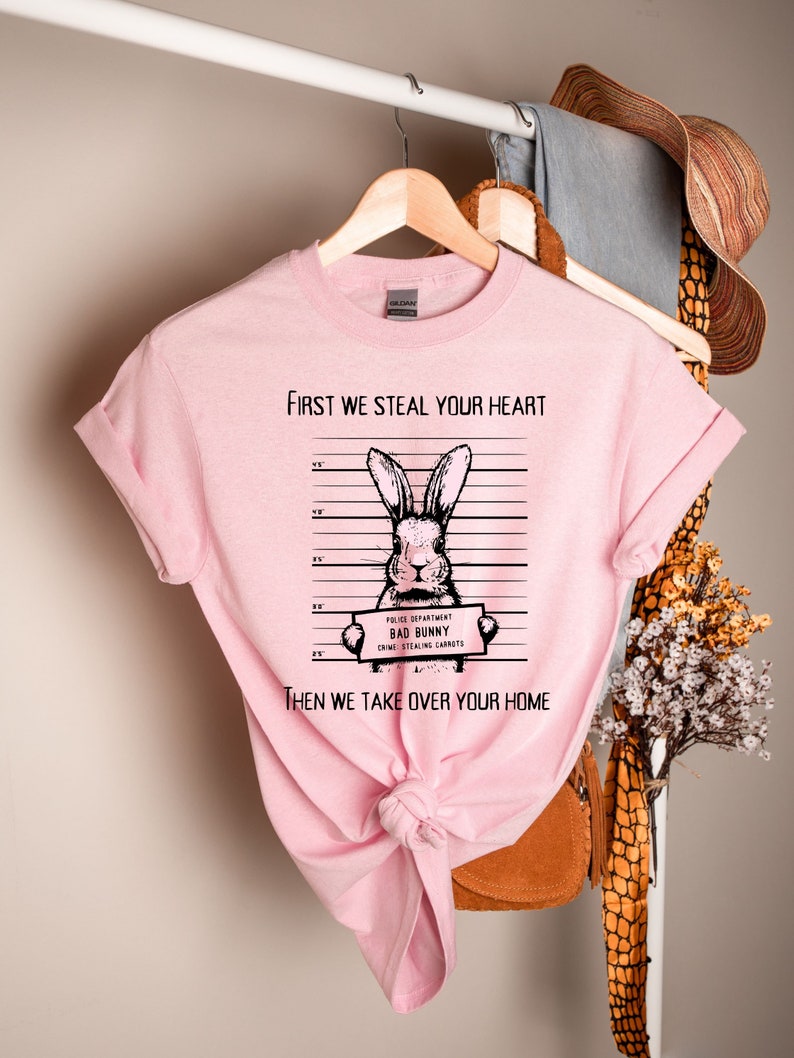 K&ouml;nnte beinhalten: Rosafarbenes T-Shirt mit einer schwarz-wei&szlig;en Illustration eines Kaninchens, das ein Schild mit der Aufschrift "Police Department Bad Bunny Crime Stealing Carrots" h&auml;lt. Der Text auf dem T-Shirt lautet "First we steal your heart then we take over your home".