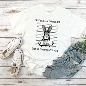 K&ouml;nnte beinhalten: Wei&szlig;es T-Shirt mit einer Schwarzwei&szlig;illustration eines Kaninchens mit dem Text "First we steal your heart" und "Then we take over your home". Das Kaninchen h&auml;lt ein Schild mit der Aufschrift "Police Department Bad Bunny Crime: Stealing Carrots".