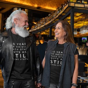 Może przedstawiać: Para w czarnych koszulkach z białym napisem "O YEAF OF DRIVII ACH OTH UTS AN TIL DING STRON" i "O YEA OF DRIVI EACH OTH NUTS A STIL COING STR". Stoją w barze i uśmiechają się do siebie.