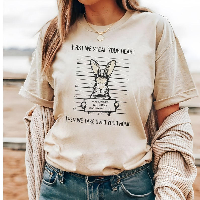 K&ouml;nnte beinhalten: Ein beiges T-Shirt mit einem Grafikdruck eines Kaninchens in einer Polizeiaufstellung. Der Text auf dem Shirt lautet "First we steal your heart" und "Then we take over your home".