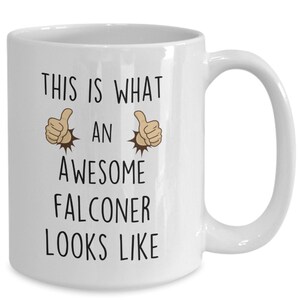 May include: White ceramic coffee mug with black text that reads "This is what an awesome falconer looks like". Two thumbs up emojis are on either side of the word "an".
