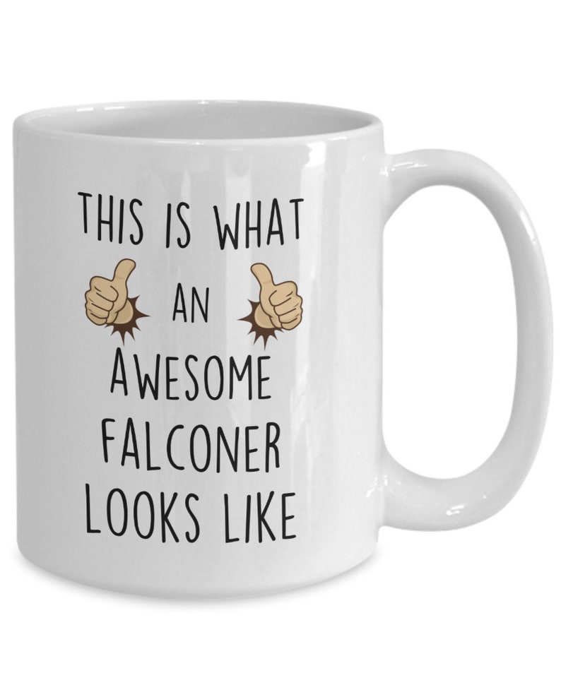 May include: White ceramic coffee mug with black text that reads "This is what an awesome falconer looks like". Two thumbs up emojis are on either side of the word "an".