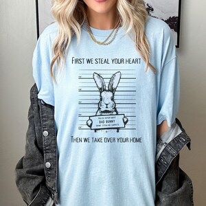 K&ouml;nnte beinhalten: Hellblaues T-Shirt mit einer schwarz-wei&szlig;en Illustration eines Kaninchens, das ein Schild mit der Aufschrift "Police Department Bad Bunny Crime: Stealing Carrots" h&auml;lt. Der Text "First we steal your heart" und "Then we take over your home" ist auf dem T-Shirt aufgedruckt.