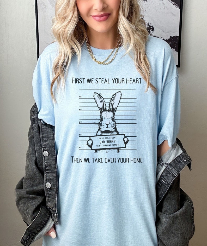 K&ouml;nnte beinhalten: Hellblaues T-Shirt mit einer schwarz-wei&szlig;en Illustration eines Kaninchens, das ein Schild mit der Aufschrift "Police Department Bad Bunny Crime: Stealing Carrots" h&auml;lt. Der Text "First we steal your heart" und "Then we take over your home" ist auf dem T-Shirt aufgedruckt.