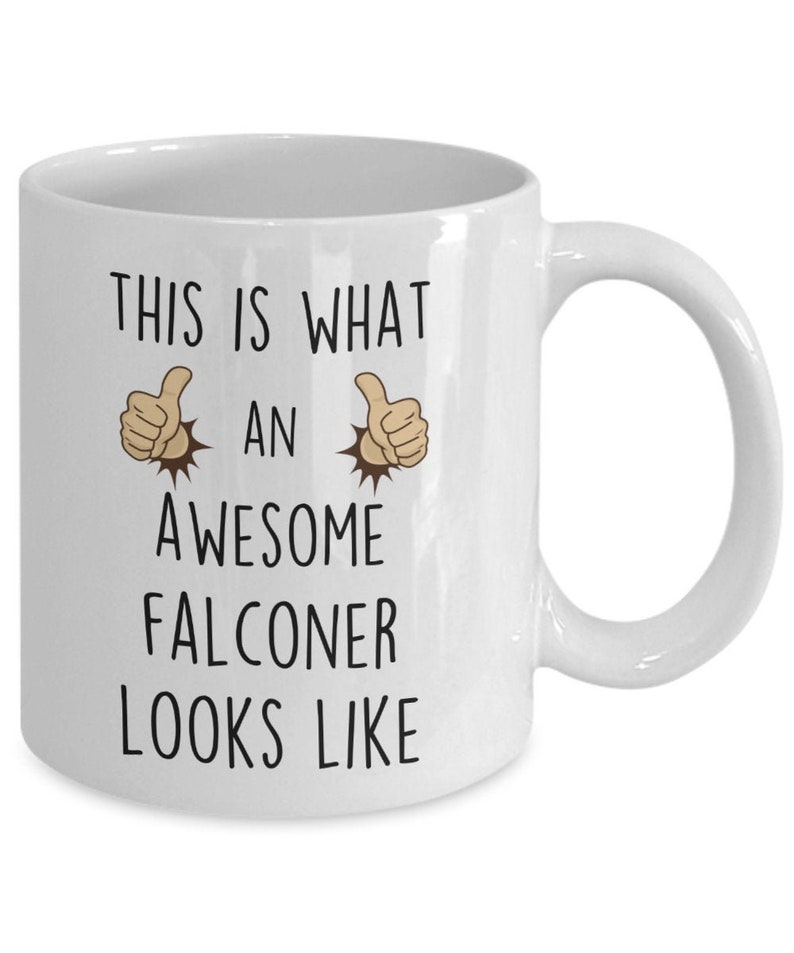 May include: White ceramic coffee mug with black text that reads "This is what an awesome falconer looks like". Two thumbs up emojis are on either side of the word "an".