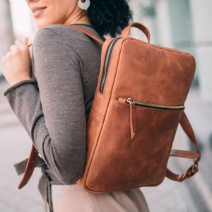 May include: A woman wearing a gray fedora and a brown leather backpack. The backpack has a zippered pocket and a flap closure.