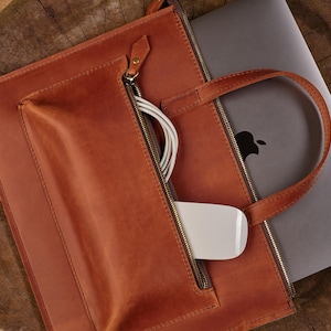 Peut inclure: Un sac d'ordinateur portable en cuir marron avec une fermeture éclair et une poignée. Le sac a une poche avant avec une fermeture éclair, contenant une souris blanche et un câble de chargement blanc. Un ordinateur portable argenté avec le logo Apple est à l'intérieur du sac.