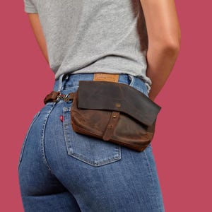 Könnte beinhalten: Eine dunkelbraune Gürteltasche aus Leder mit Umschlagklappe, einem einzelnen Metalldruckknopf und einem dünnen Lederriemen. Die Tasche ist an einem Metallring befestigt und wird über einer blauen Jeans und einem grauen T-Shirt getragen.